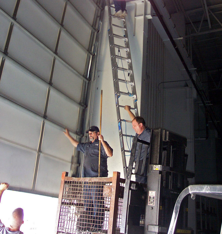 Two guys standing in a parts basket, hoisted up by a hi-lo, with and a guy at the top of the ladder.